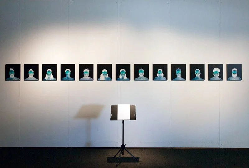 A series of framed photographs of a person wearing a medical mask and glasses, displayed on a white gallery wall. A music stand is positioned in front of the photos.