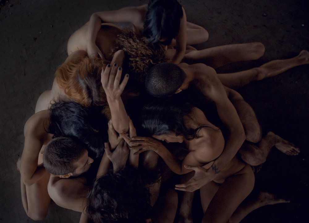 Group of naked people intertwined in a pile on a dark wooden floor.