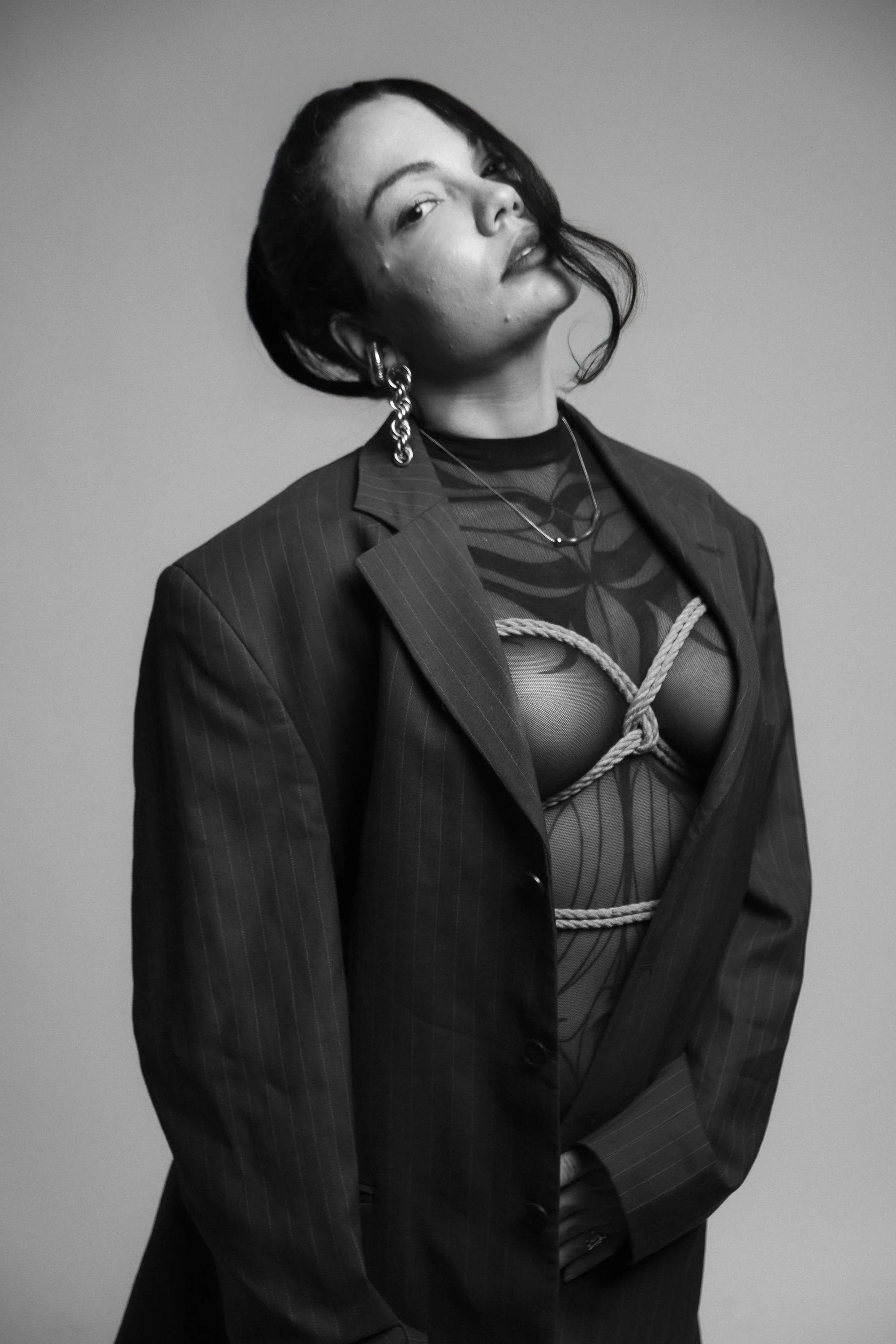 Black and white photo of a woman with short hair wearing a pinstripe blazer, mesh top with decorative rope, multiple earrings, and layered necklaces, posing with her head tilted back slightly.
