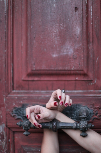 Person's hands with red painted nails and a ring, resting on a black metal latch against a weathered red wooden door.
