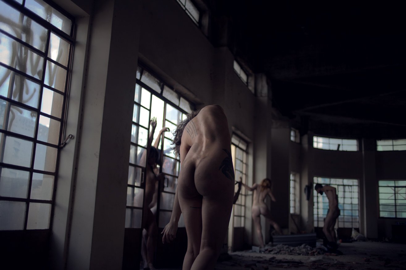 Nude individuals in an abandoned industrial building with large windows, some pressed against the windows, others standing or bending, in a dimly lit setting.