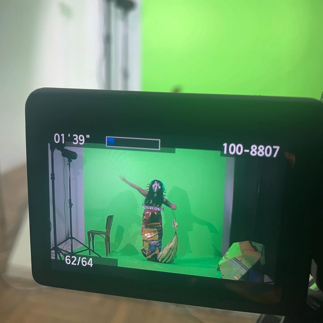 Camera screen showing a green screen with a person dressed as a traditional dancer, standing with one arm raised and holding a fan, a chair and lighting equipment nearby.