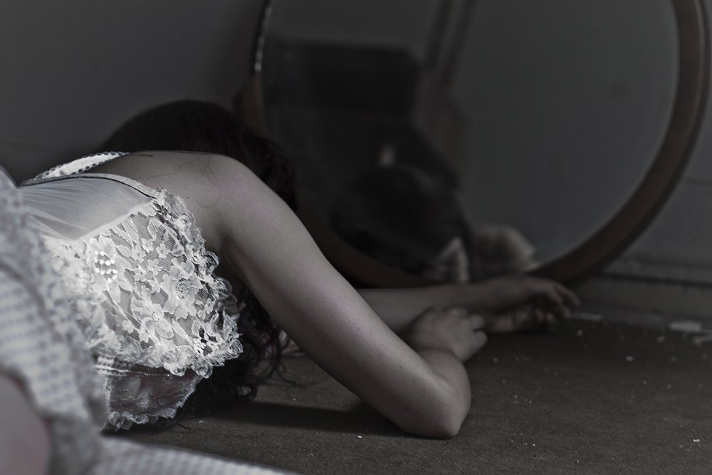 A person in a white lace dress lying on the floor with their head beside a mirror, appearing distressed or exhausted.
