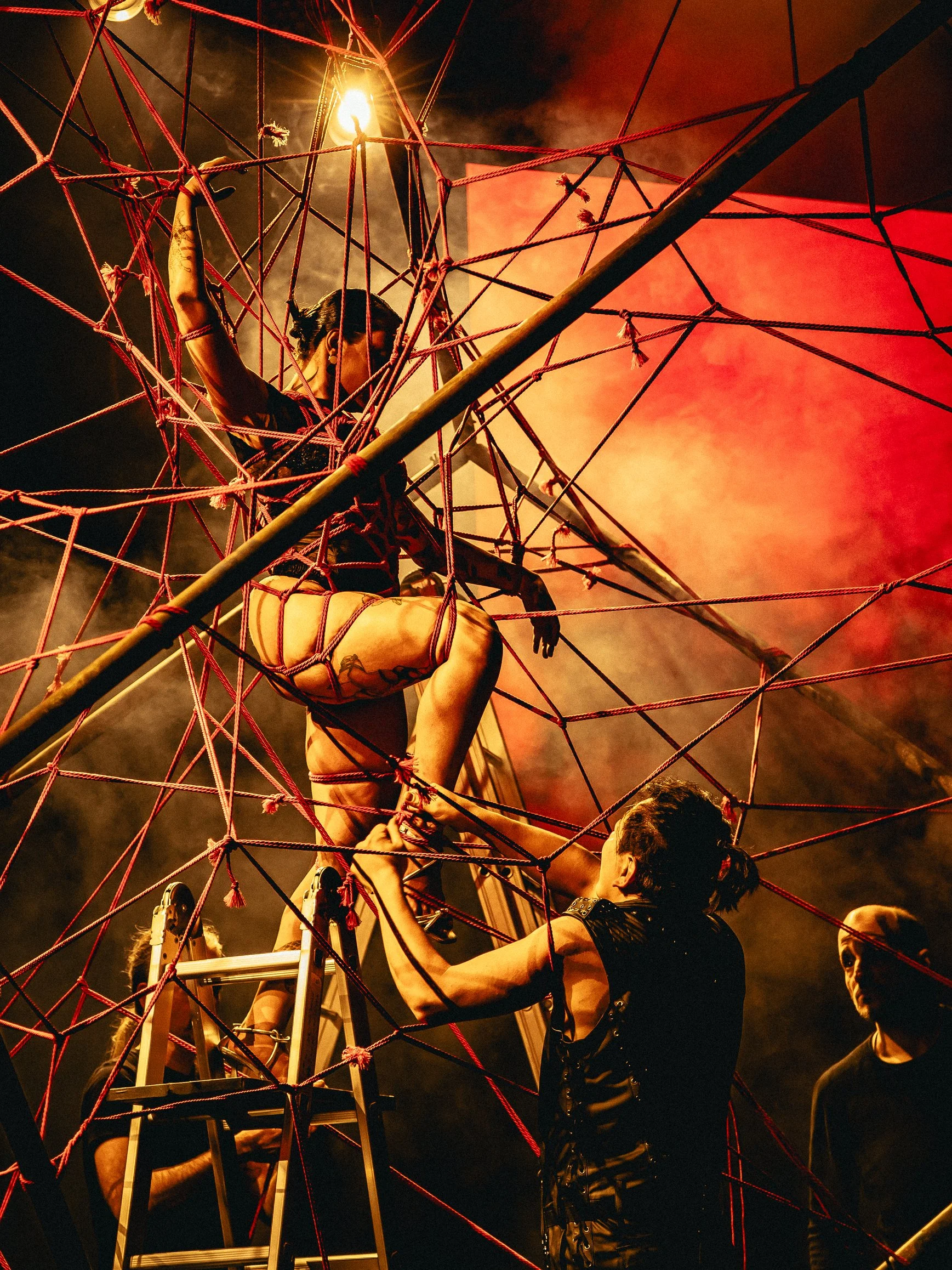Performance art scene with performers entangled in red ropes on a stage with dramatic lighting and smoke effects.
