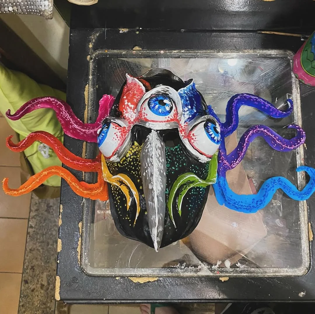 Colorful decorative mask with multiple painted blue eyes and multicolored tentacle-like appendages.