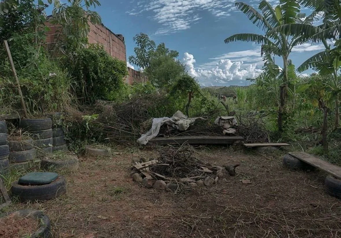 An outdoor backyard space with a circle of stones around a small fire pit, surrounded by stacked tires and makeshift benches, with trees and bushes in the background under a partly cloudy sky.