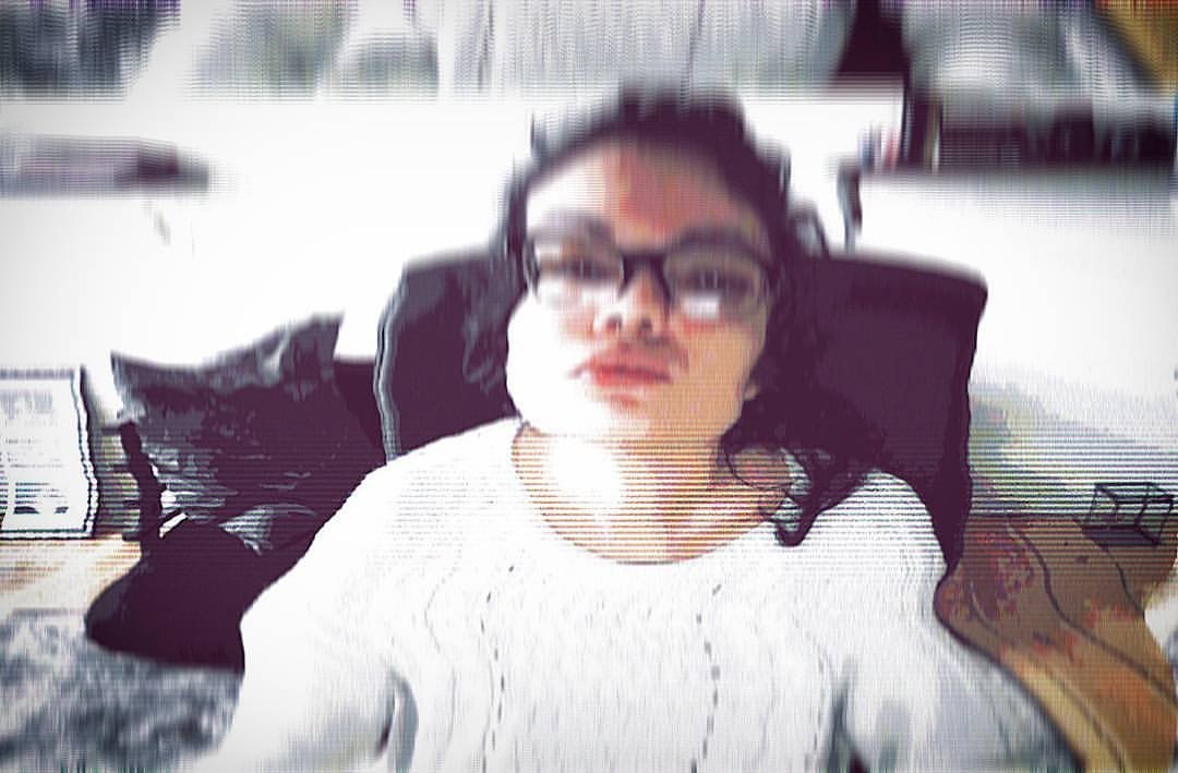 A woman with dark curly hair and glasses, sitting in a wheelchair, in a room with a desk and various objects in the background.