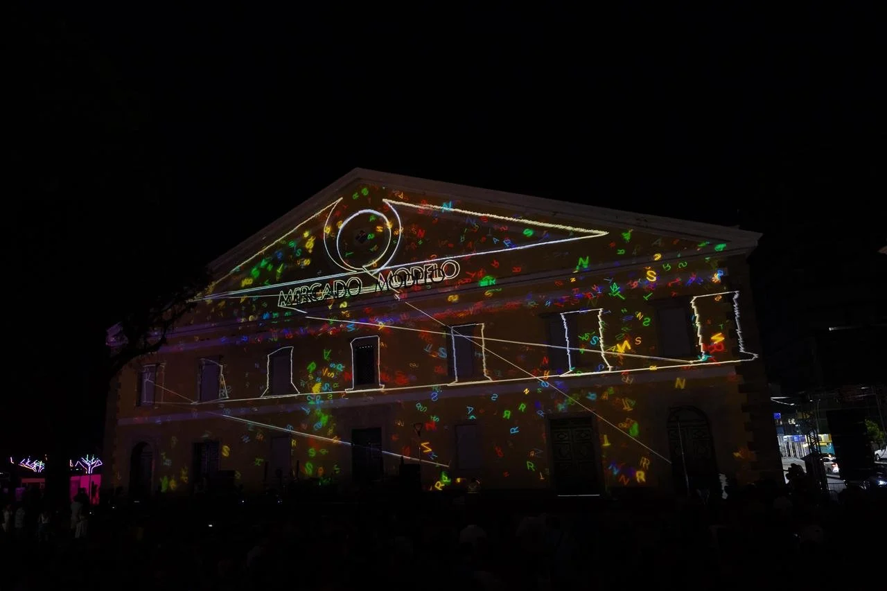 Night scene of a historic building with a light projection display, featuring colorful letters and lines, and the sign 'Museo Museo' on the facade.