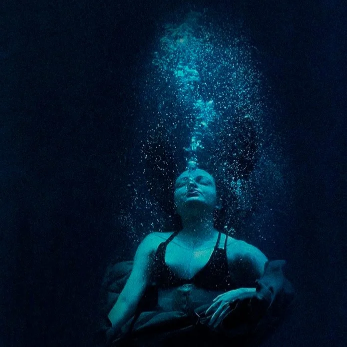 A woman underwater with closed eyes, wearing a black bikini top, with bubbles rising from her mouth.
