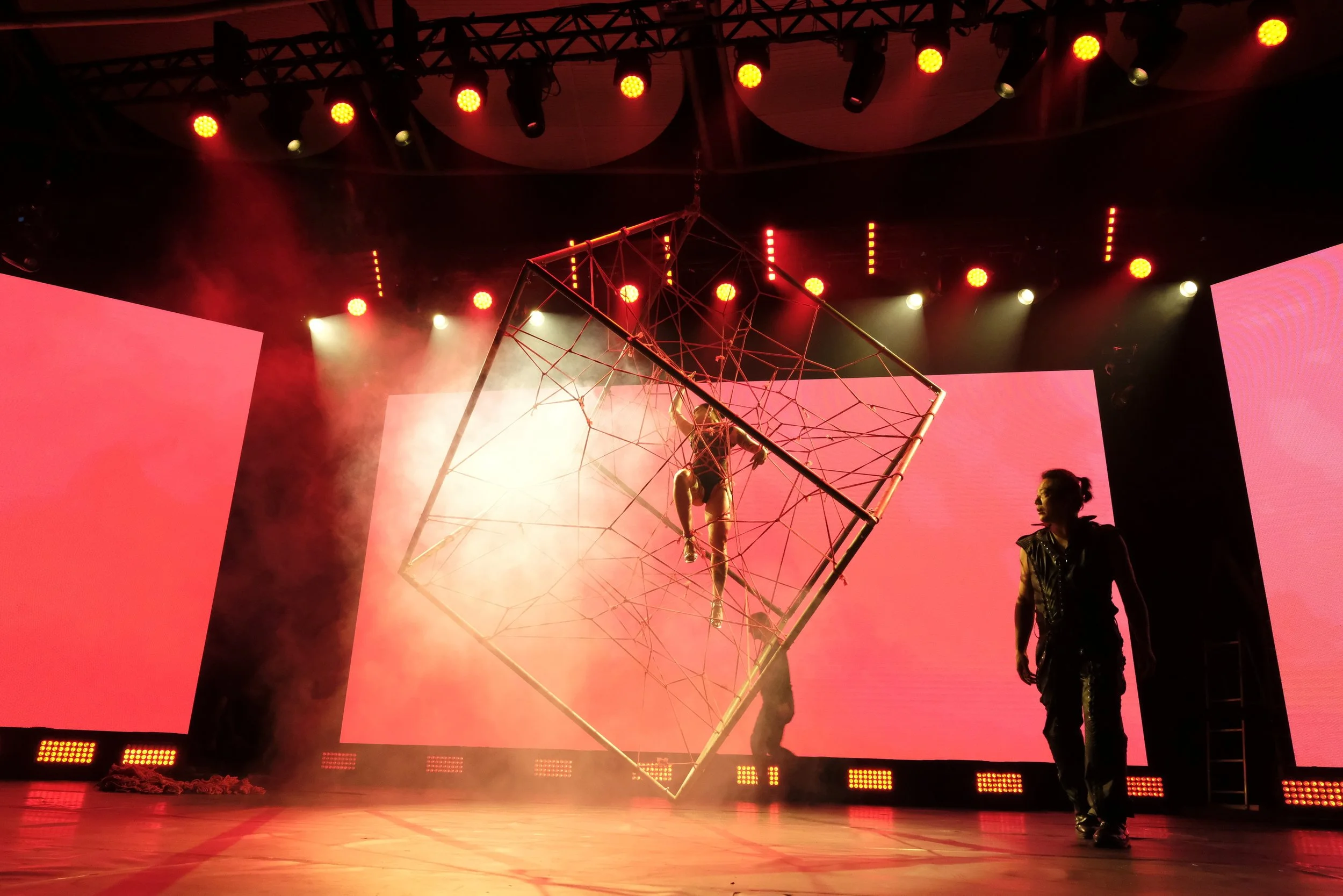Performer hanging inside a geometric wireframe cube on stage with red lighting and large screens in the background.