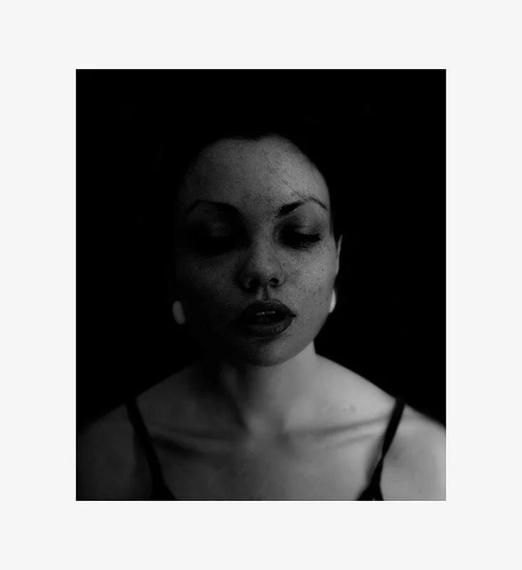 Black and white portrait of a woman with closed eyes, dark lipstick, earrings, and shoulder straps, against a black background.