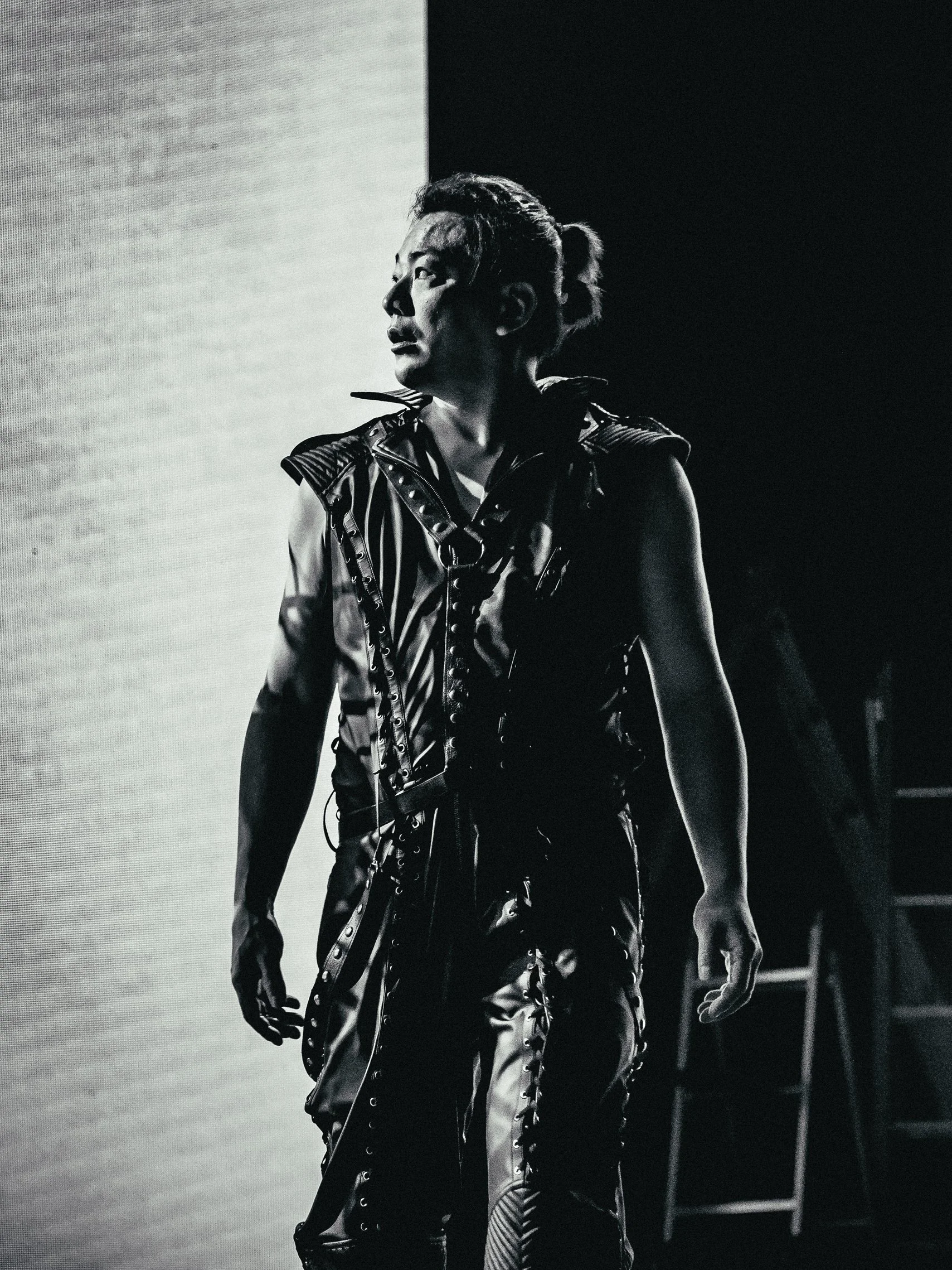 A person with short hair, wearing a sleeveless leather vest with studs, walking on stage with dramatic lighting, black and white photograph.