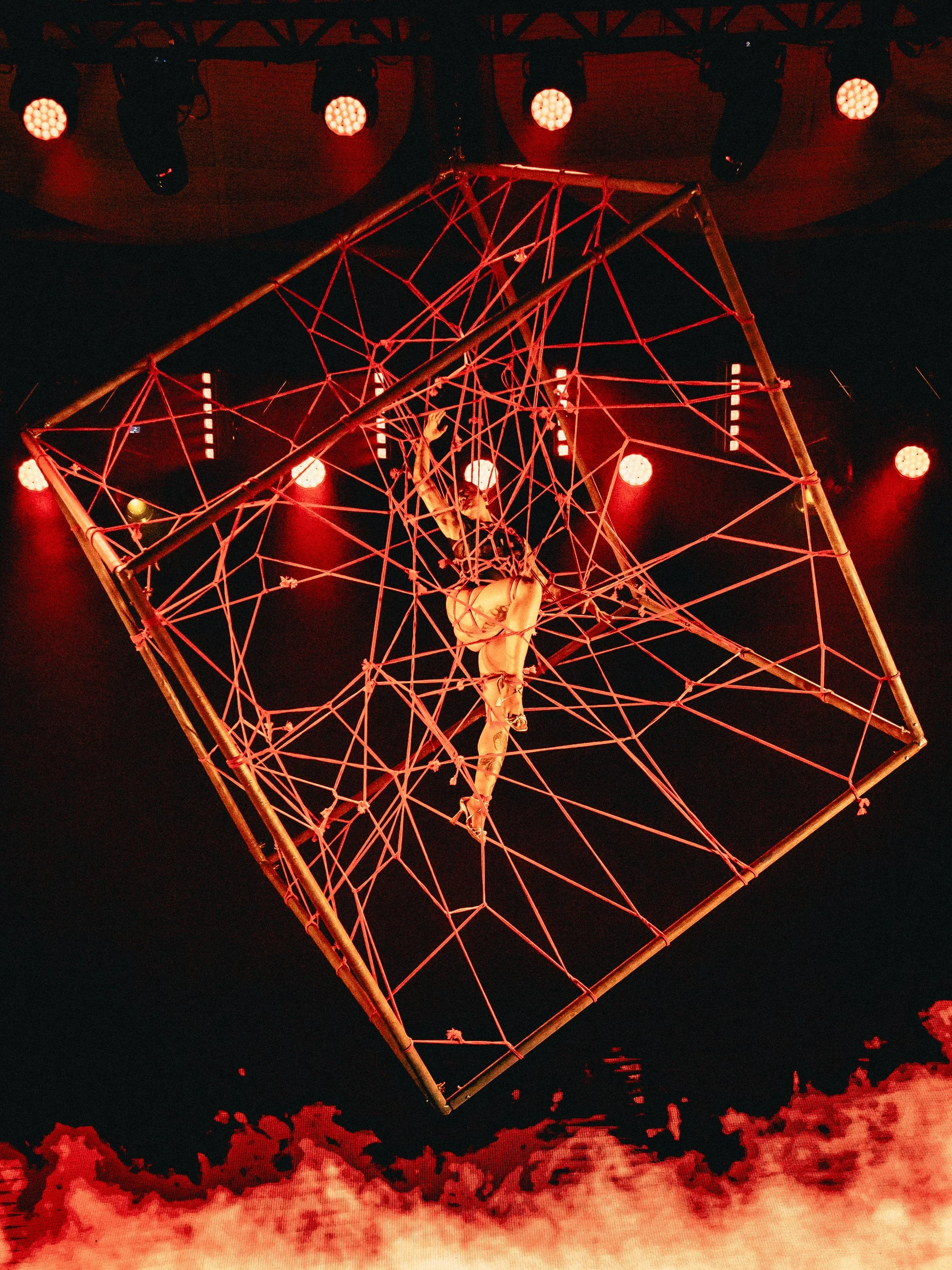 A performer hangs upside down inside a geometric metal framework during a circus or acrobatic show, illuminated by red stage lights.