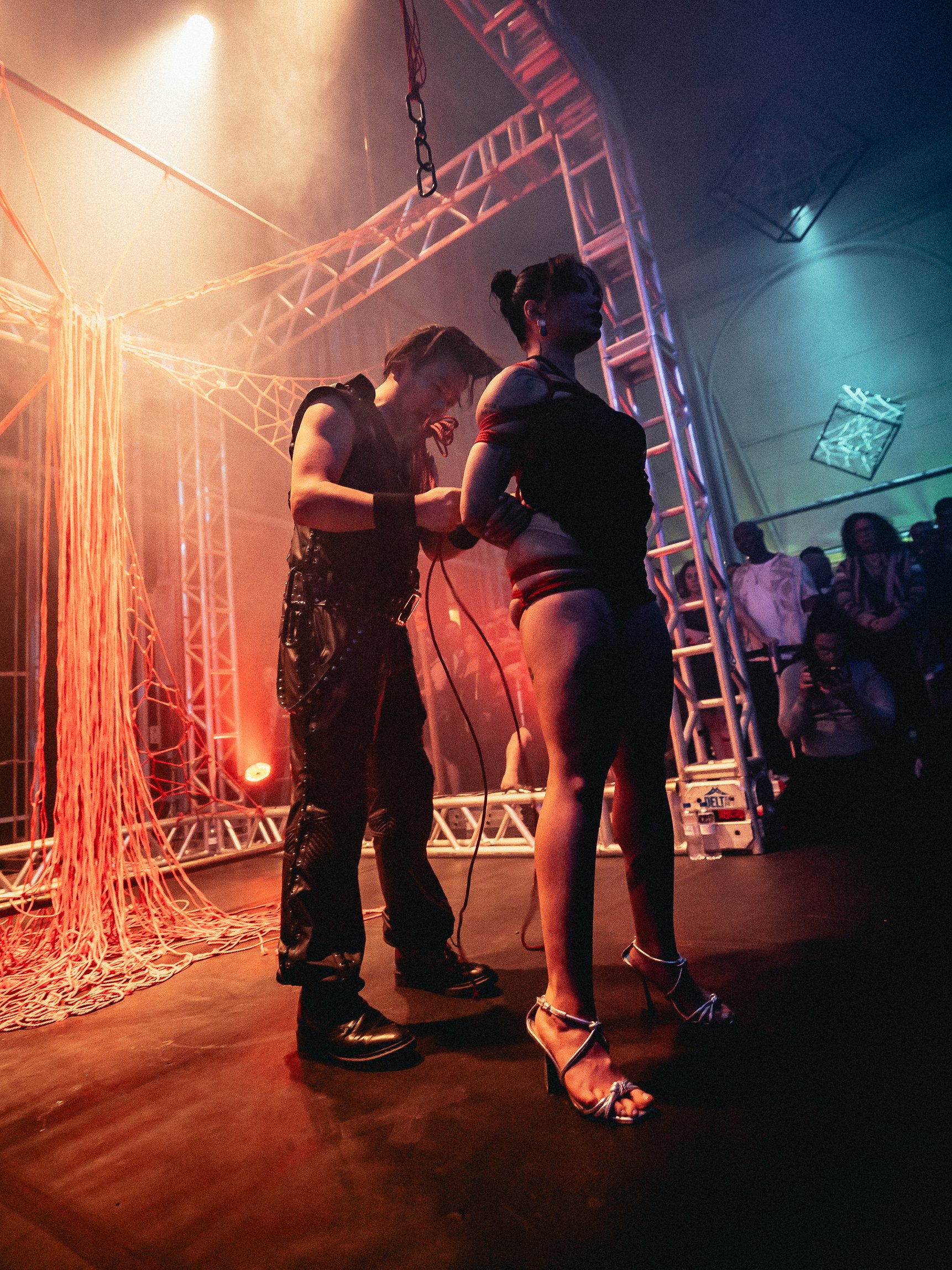 A woman on stage has her hands tied behind her back while another woman stands behind her, holding a power cord. The scene is lit with orange and blue stage lights, with audience members visible in the background. The stage has metal trusses and red 