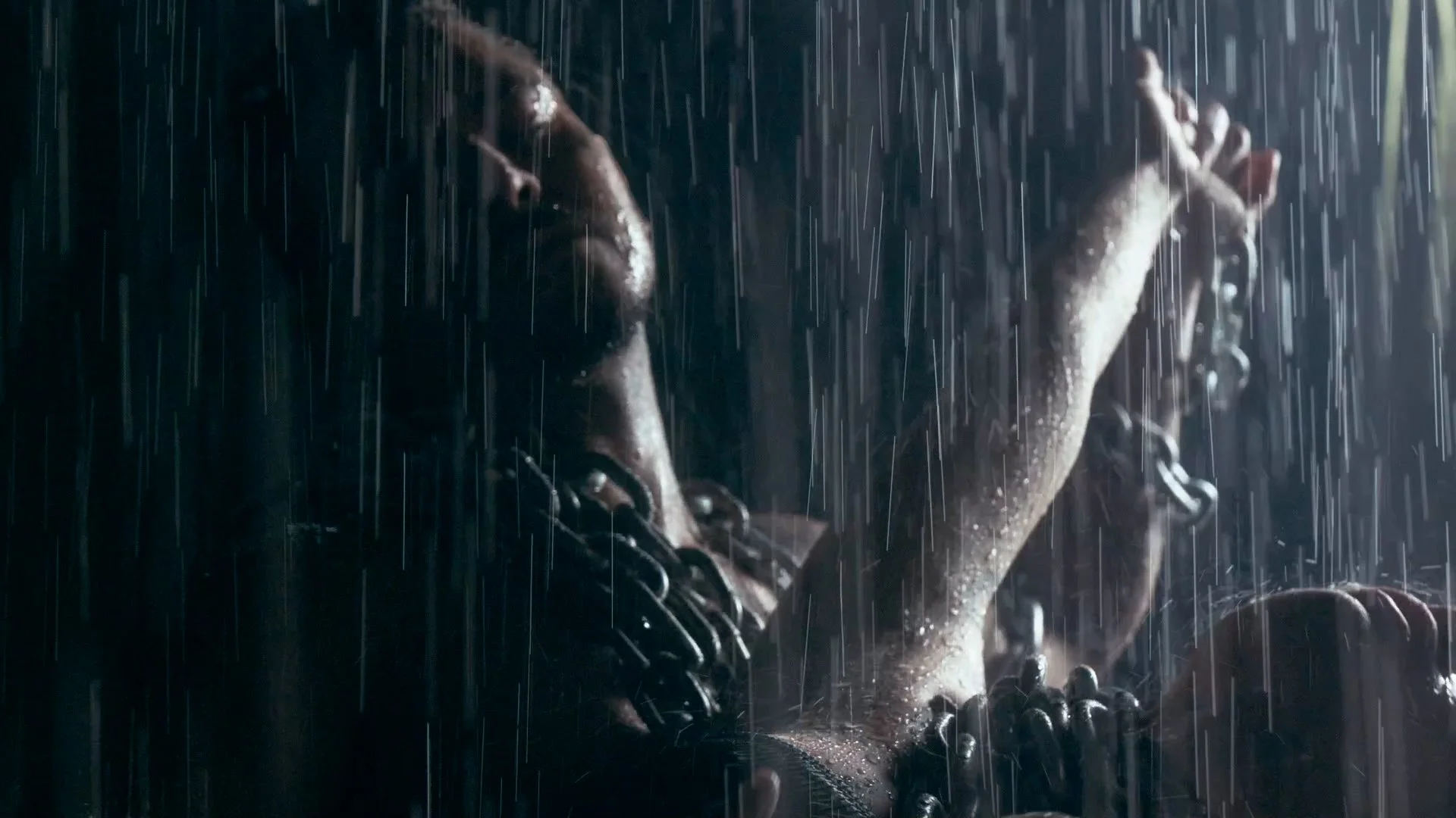 A woman with chains around her wrists and neck, standing in the rain with her eyes closed, appearing to be in distress or deep thought.