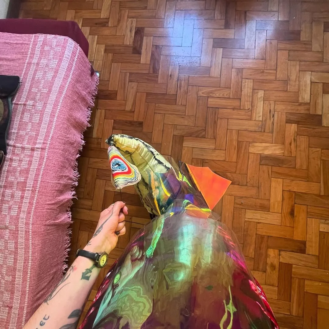 Hand holding a small colorful pinwheel, standing on a wooden floor next to a pink rug, with part of a red couch visible on the left.