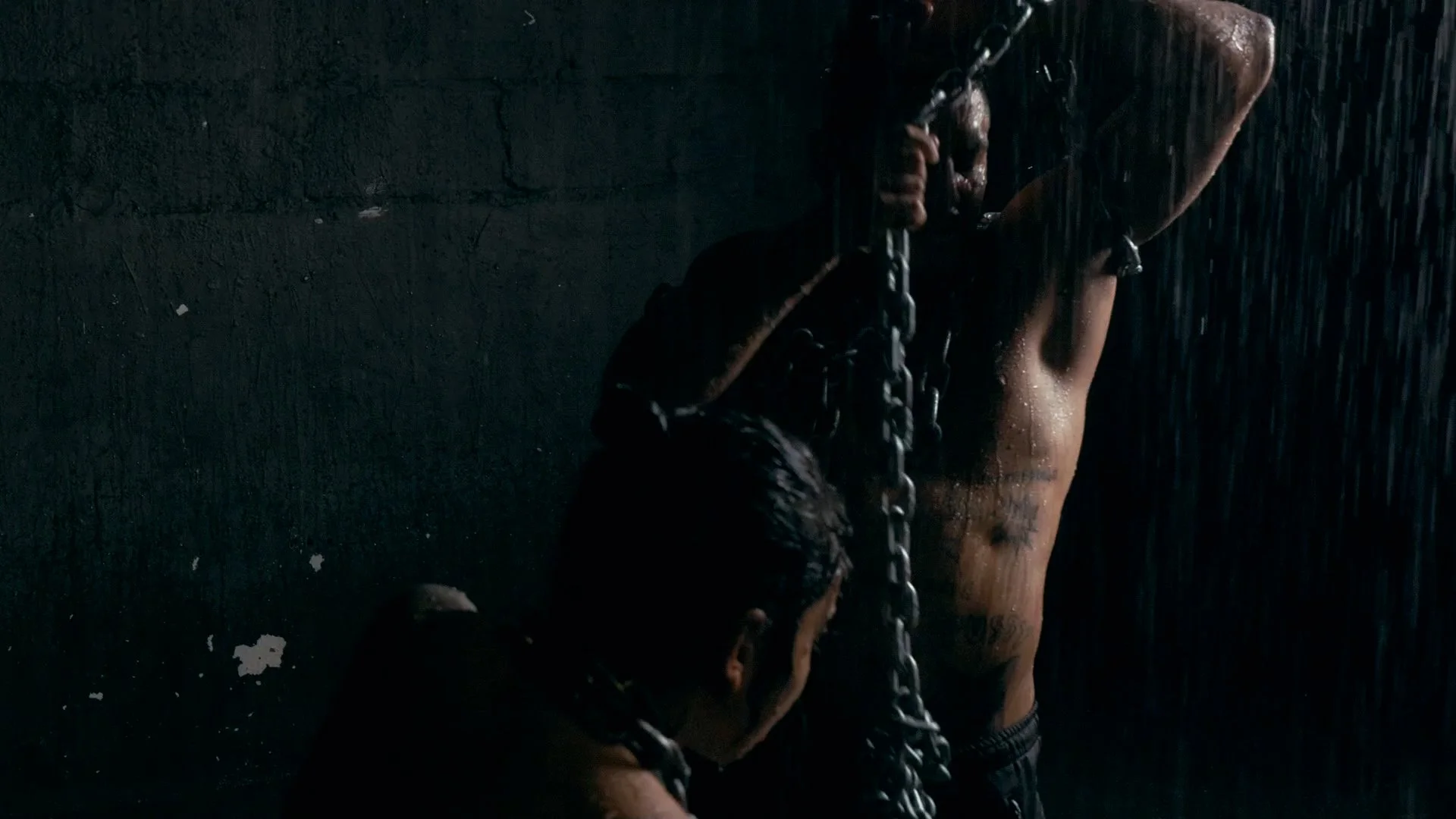 A shirtless man with tattoos on his torso is standing under heavy rain, holding a chain with both hands, with his face partially obscured.