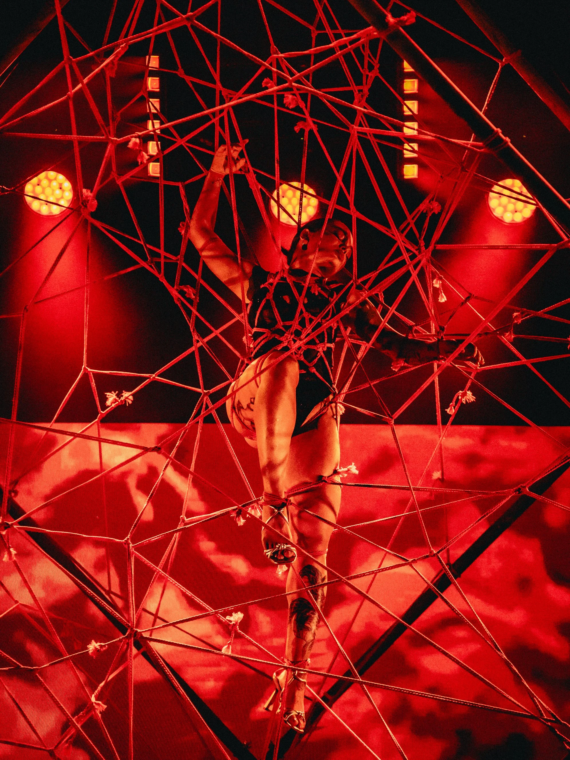 A performer hanging upside down inside a red geometric metal structure on a dark stage, illuminated by red lights.