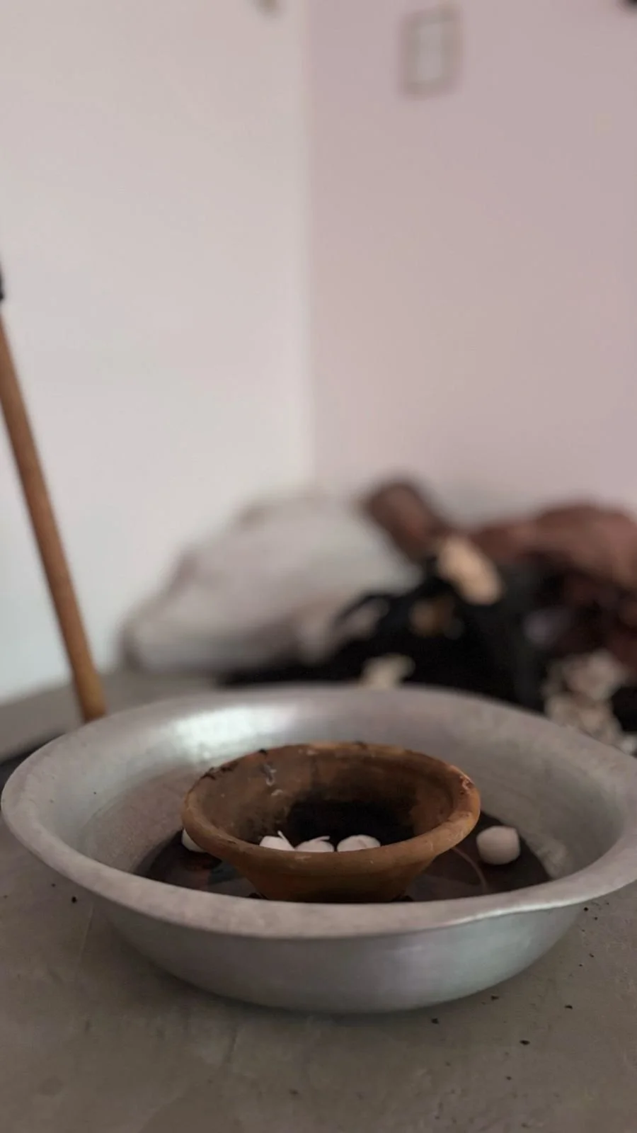A traditional oil lamp with a clay wick holder and white pebbles inside a metal basin, with a blurred background of bedding and a wall.