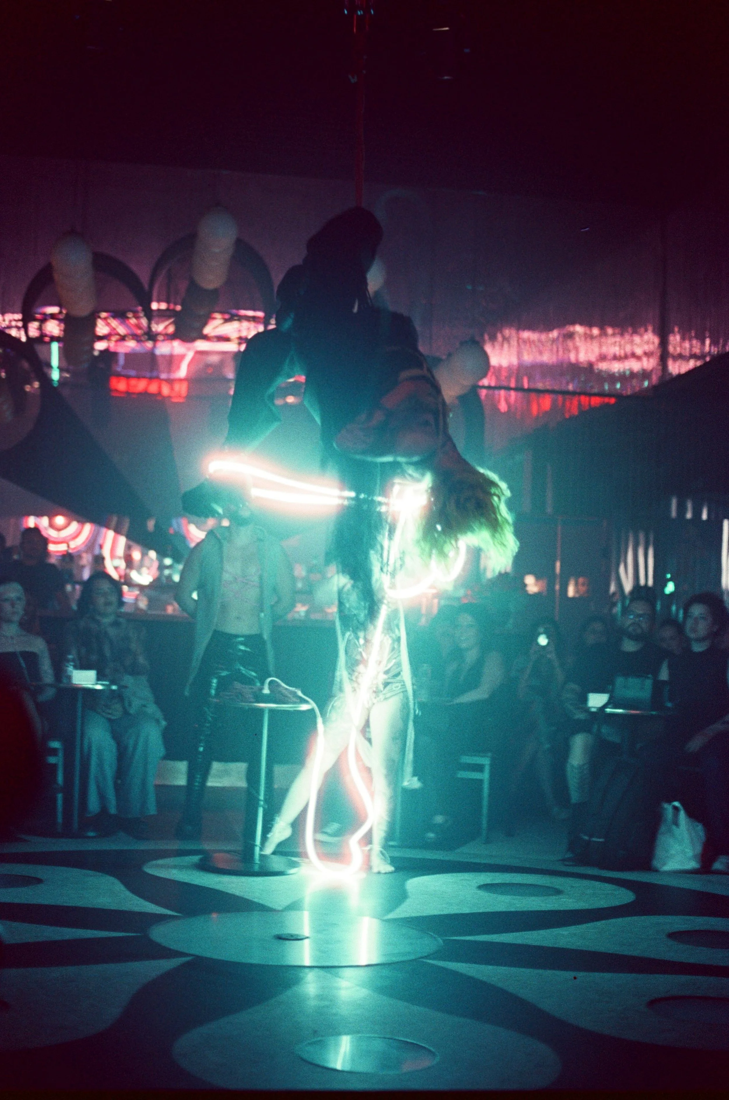 A performer on a pole with light streaks dancing in front of an audience in a nightclub or entertainment venue.
