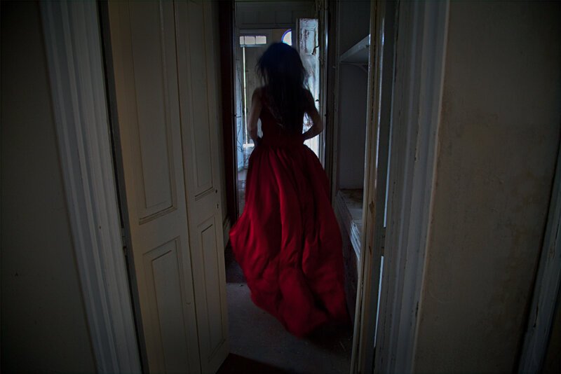 A woman in a long red dress walking away from the camera in a narrow, rustic hallway.