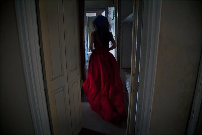 A woman in a long red dress walking through a doorway in a dimly lit, old house.