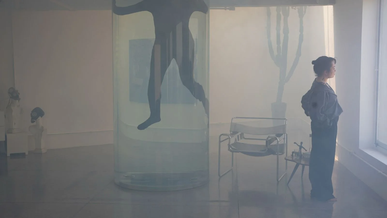 An art gallery with a woman standing near a window, reflecting her silhouette and a cactus painting on the wall, with sculptures on display.