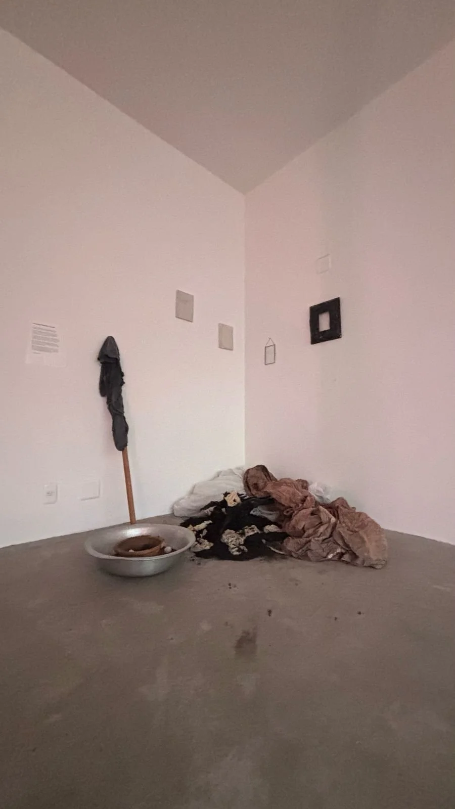 Corner of a room with plain white walls, cluttered with a mop, in a metal basin, a pile of blankets or clothes on the floor, and unorganized items. Wall decorations include three small frames and one hanging mirror or frame.
