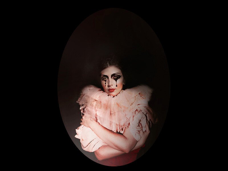 A woman with heavy, artistic makeup and tears painted on her face, wearing a ruffled, light-colored costume, inside a dark oval frame.
