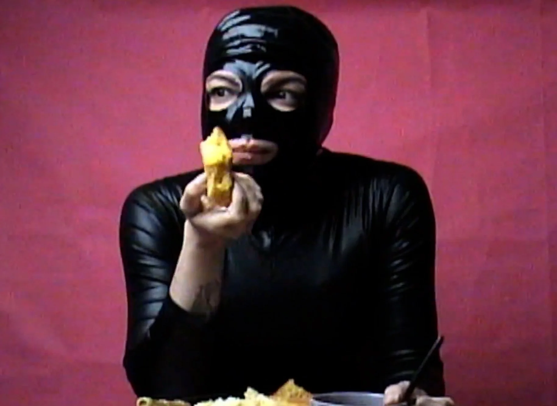 Person wearing a black mask and black leather outfit, holding and eating a yellow ice cream cone against a pink background.