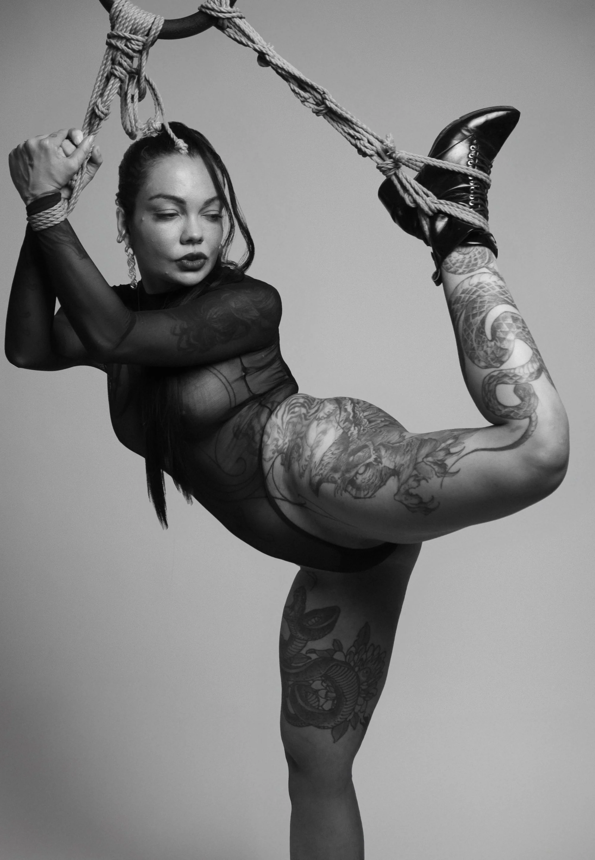 A woman with tattoos in minimal clothing poses with her leg raised, holding onto a rope and wearing shiny black boots. The image is in black and white.
