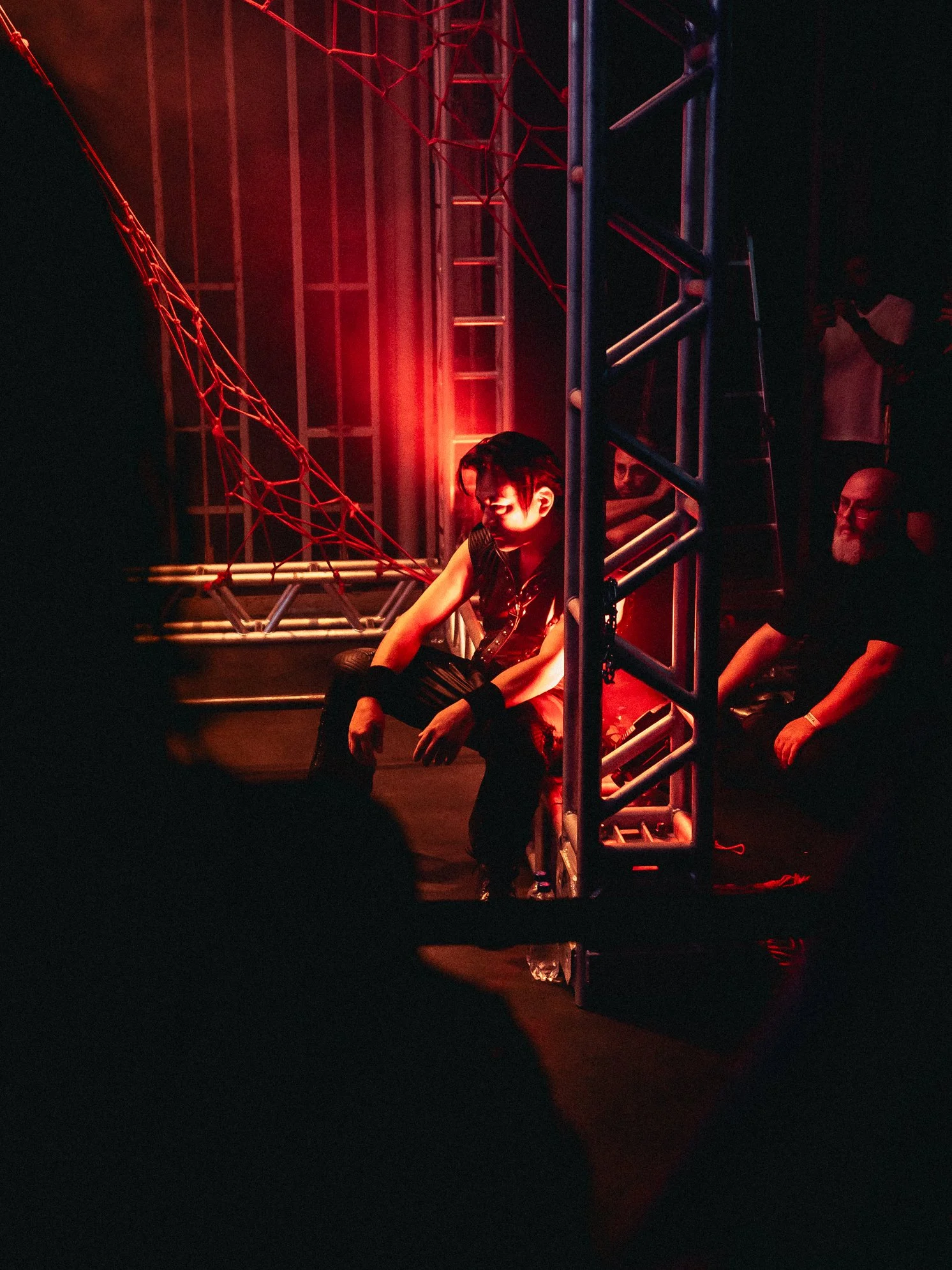 Person sitting on the ground in a dimly lit setting with red lighting, surrounded by metal scaffolding and netting, with other people in the background.