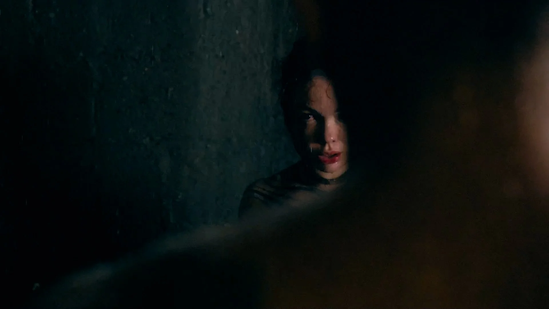 A person with wet hair and smeared makeup looks distressed, partially illuminated in a dark environment, with shadows and dim lighting on their face and background.