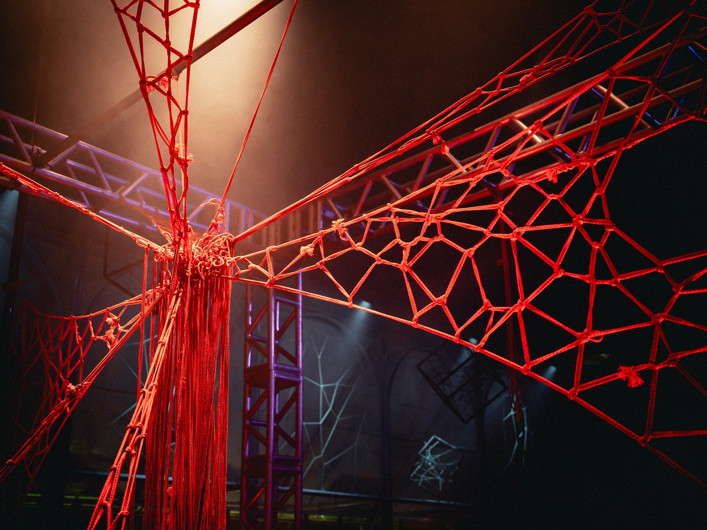 Close-up of an intricate red web structure made of ropes, with industrial metal framework and faint blue and purple lighting in the background, under a dark sky.