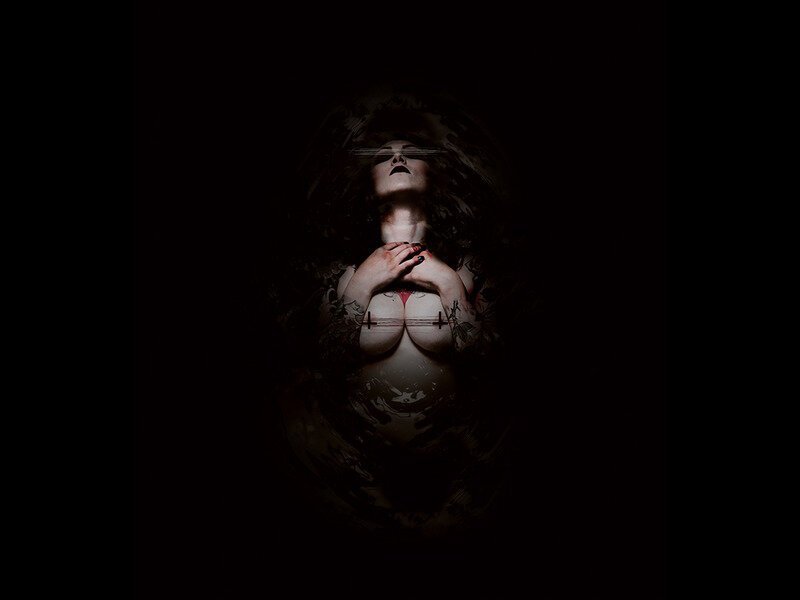 A woman with dark curly hair and tattoos on her arms and chest, holding her hands near her neck, in a dark, moody setting with dramatic lighting.
