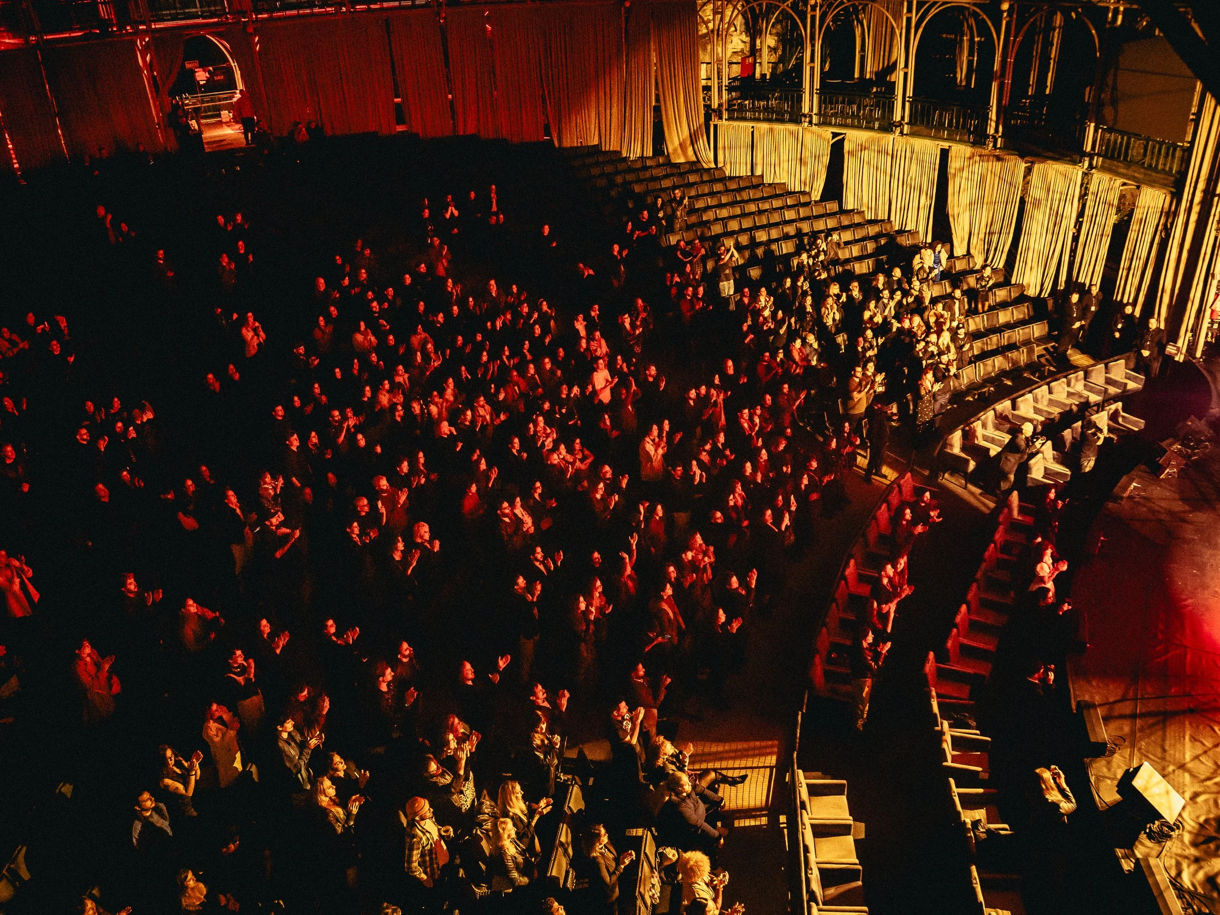 Audience seated in an auditorium, illuminated by warm, orange stage lighting, with some standing or clapping.