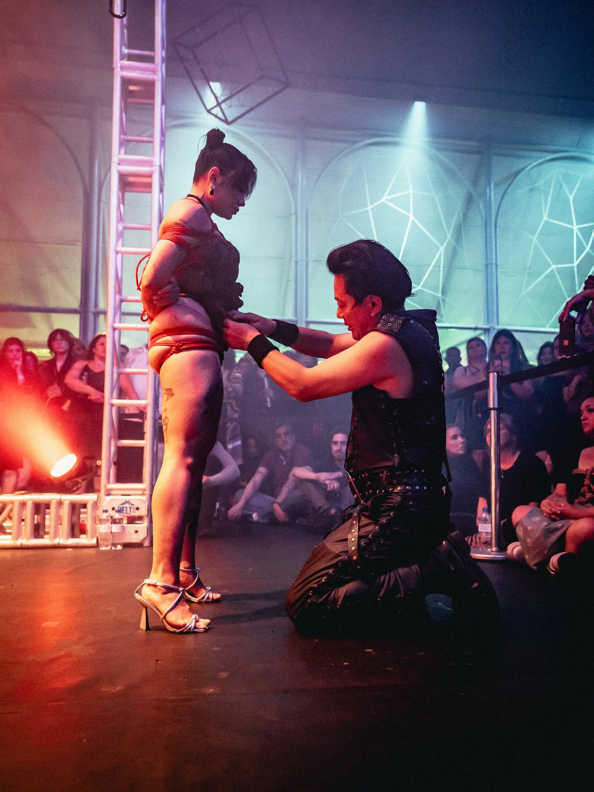 Person dressed in black kneeling and attaching a safety harness to a woman standing in a black outfit, with an audience watching in a dark, industrial-style setting with colorful lighting.