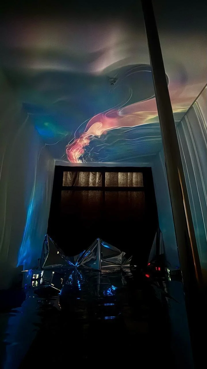 Room with a window covered by a dark curtain, colorful projected light patterns on the ceiling, and reflective metallic fabric on the floor creating an abstract, futuristic ambiance.
