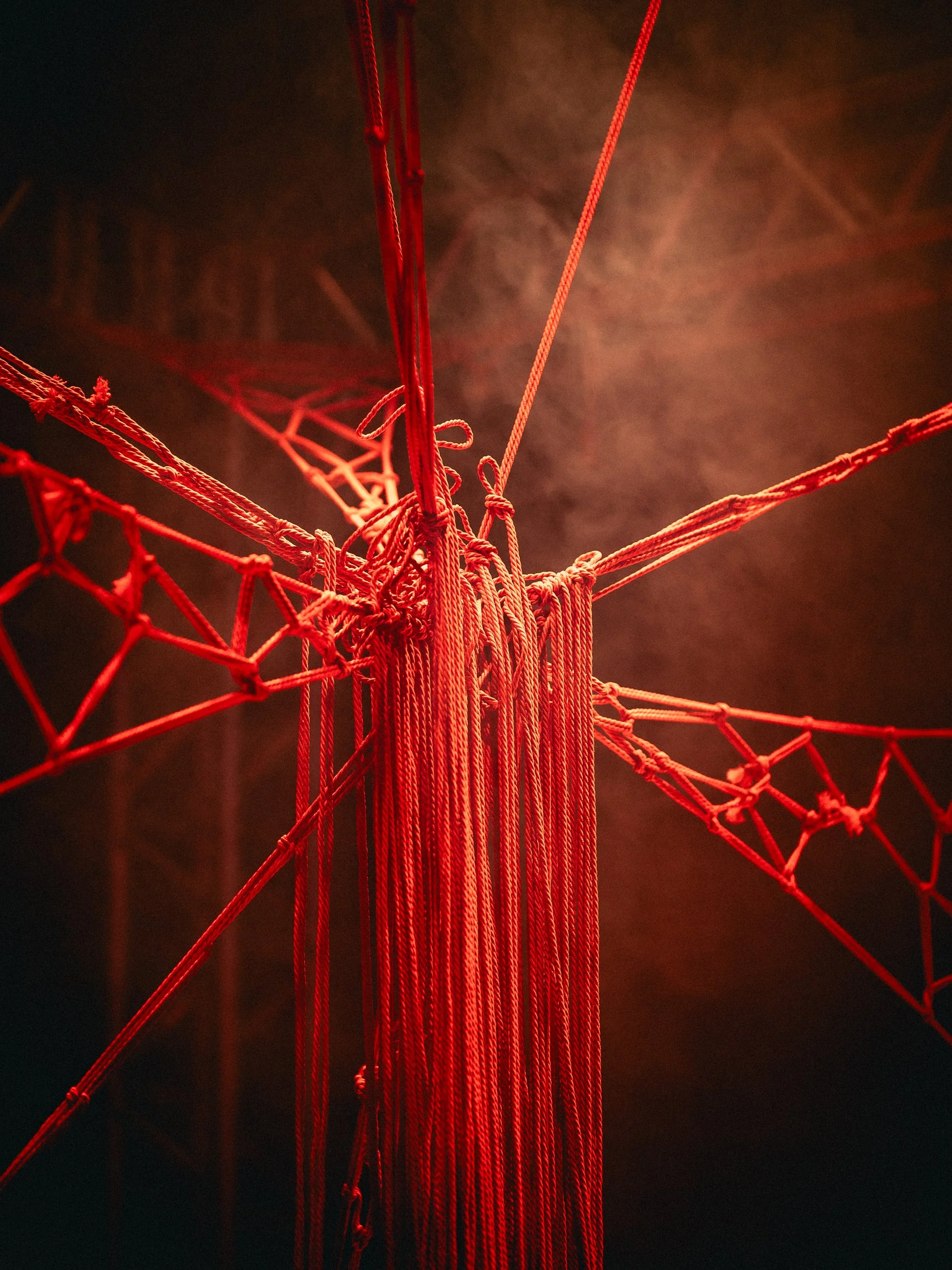 Close-up of a red string art installation with intricate woven knots and strings extending outward, illuminated with a reddish glow.