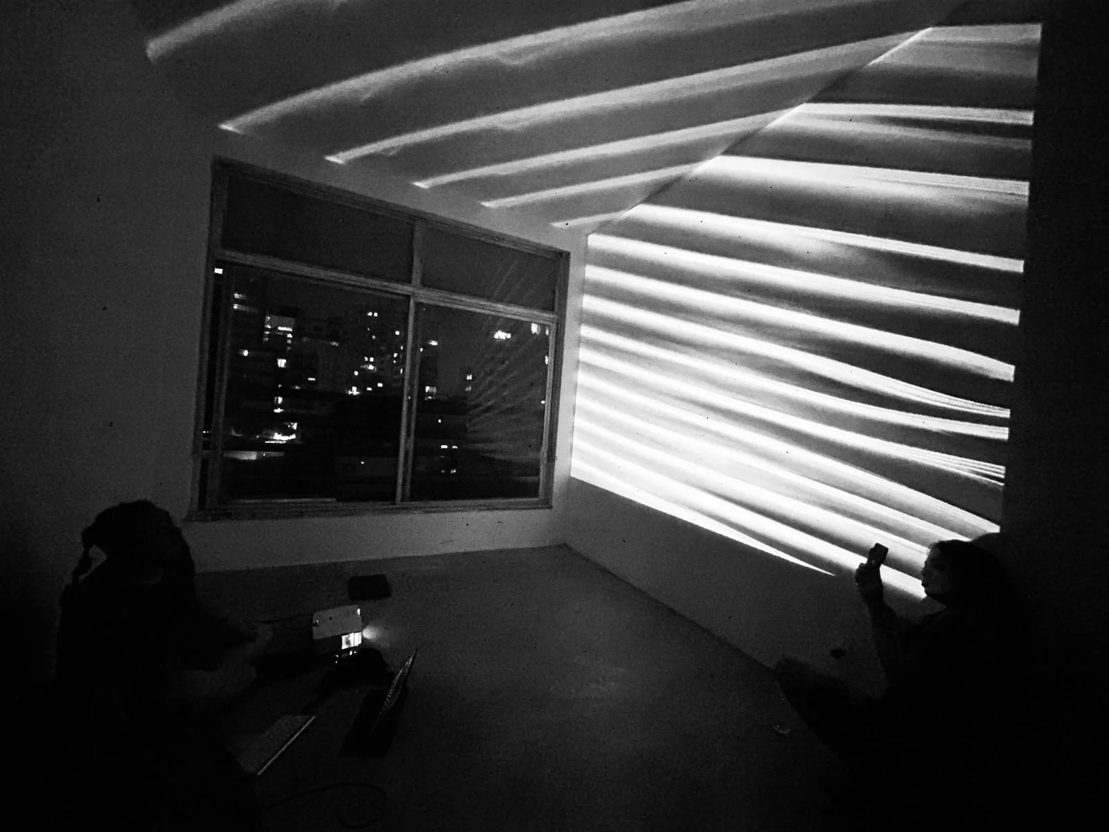 Black and white photo of two people in a room with decorative lighting on the wall and a large window showing a cityscape at night.