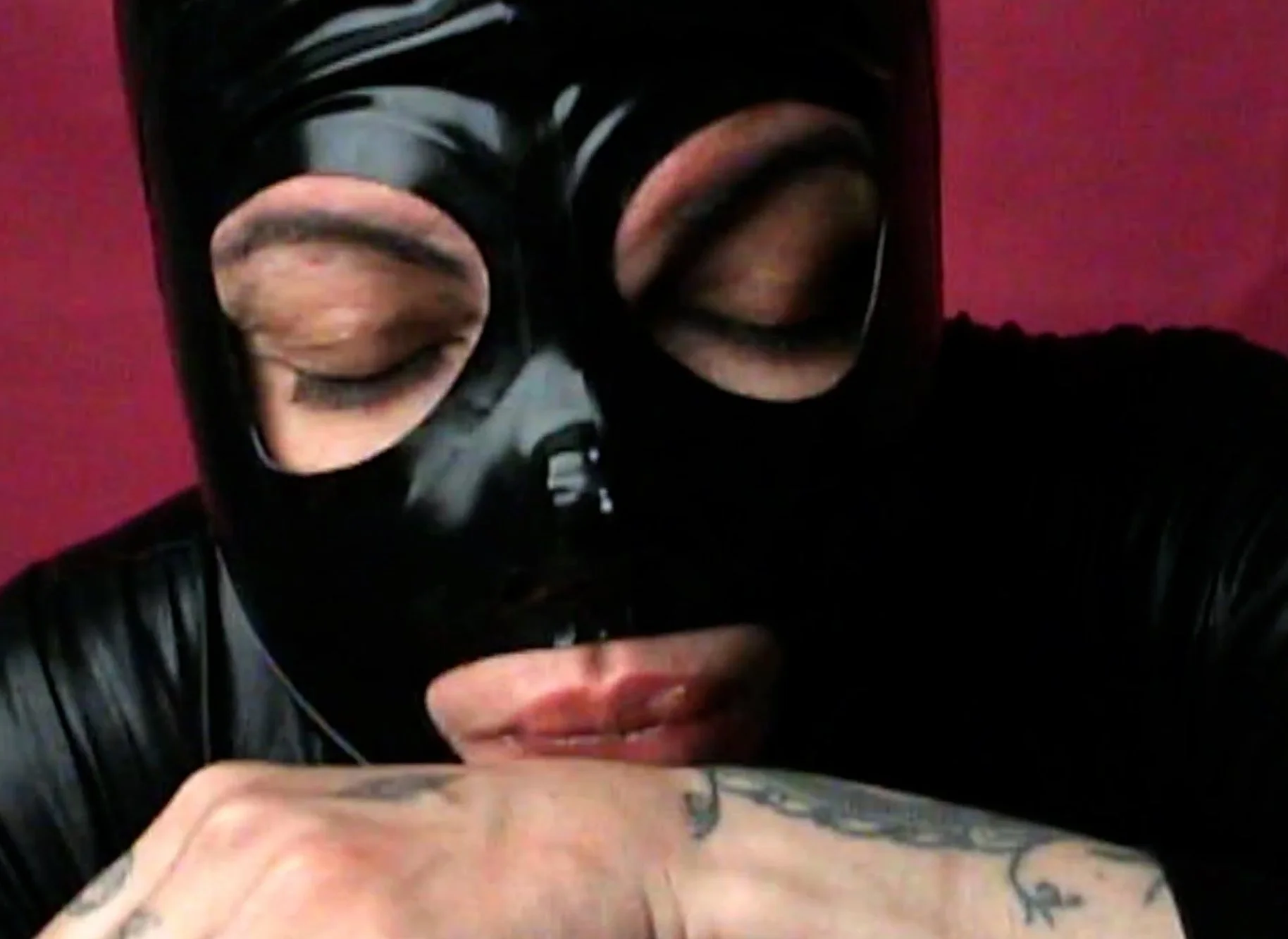 Person wearing a black mask with reflective eye lenses and a black leather jacket, covering their face, with tattoos visible on their arm, against a red background.