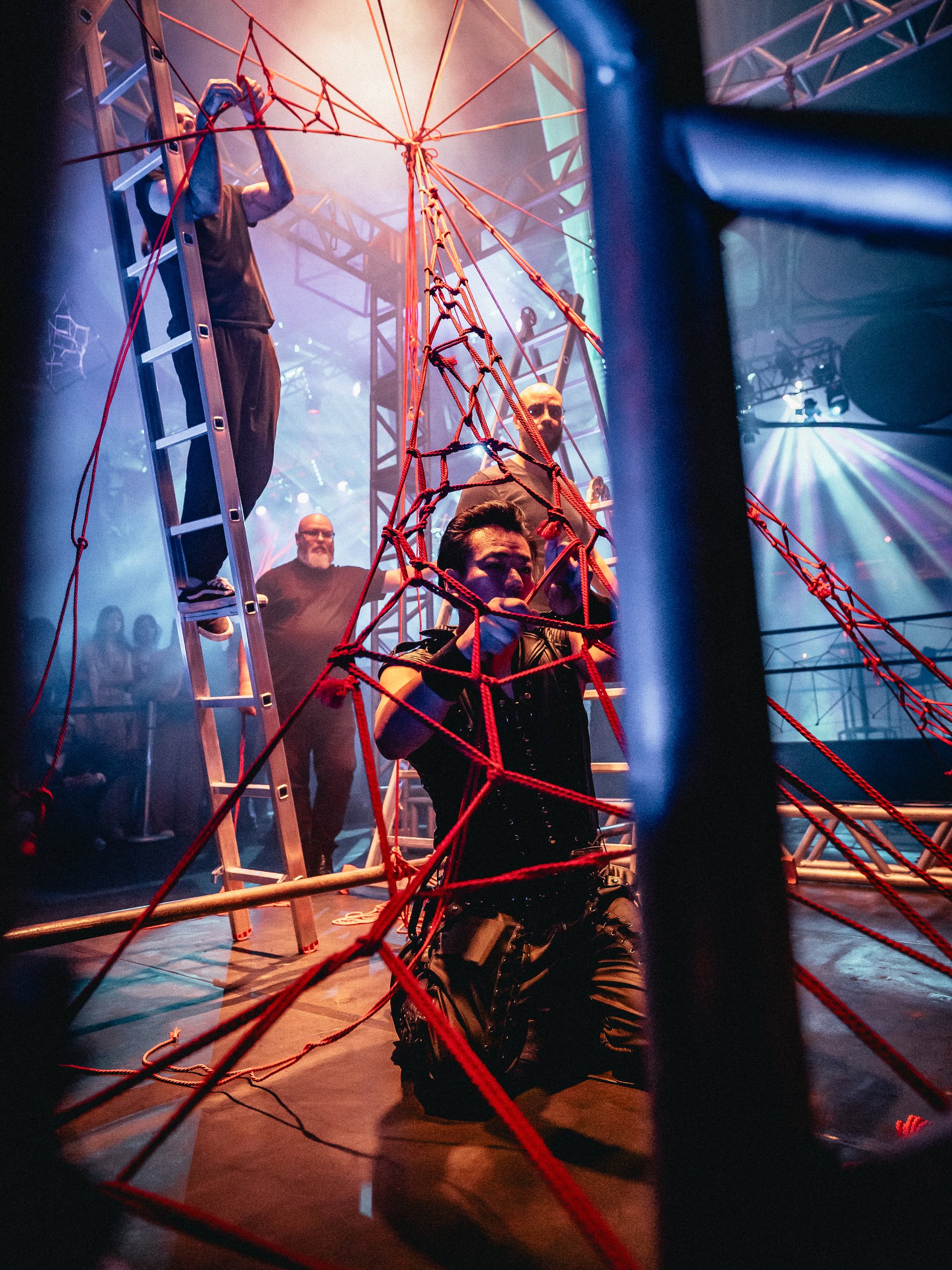 People setting up a theatrical scene with red ropes and a ladder in a dimly lit venue with stage lighting.