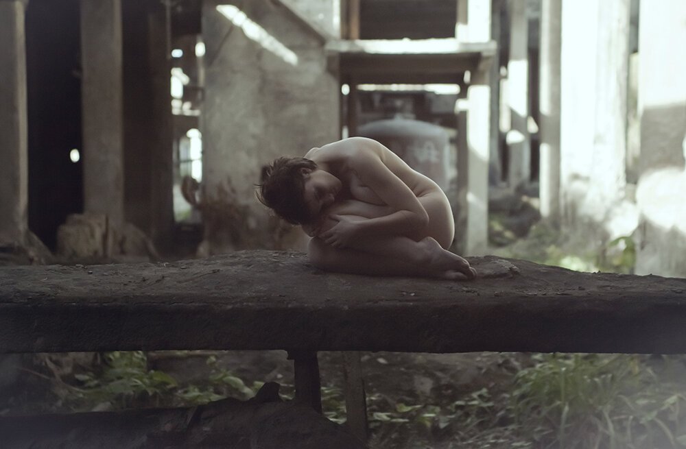 A person curled up in a fetal position, sitting on a large piece of wood in a dimly lit, abandoned or rustic setting.