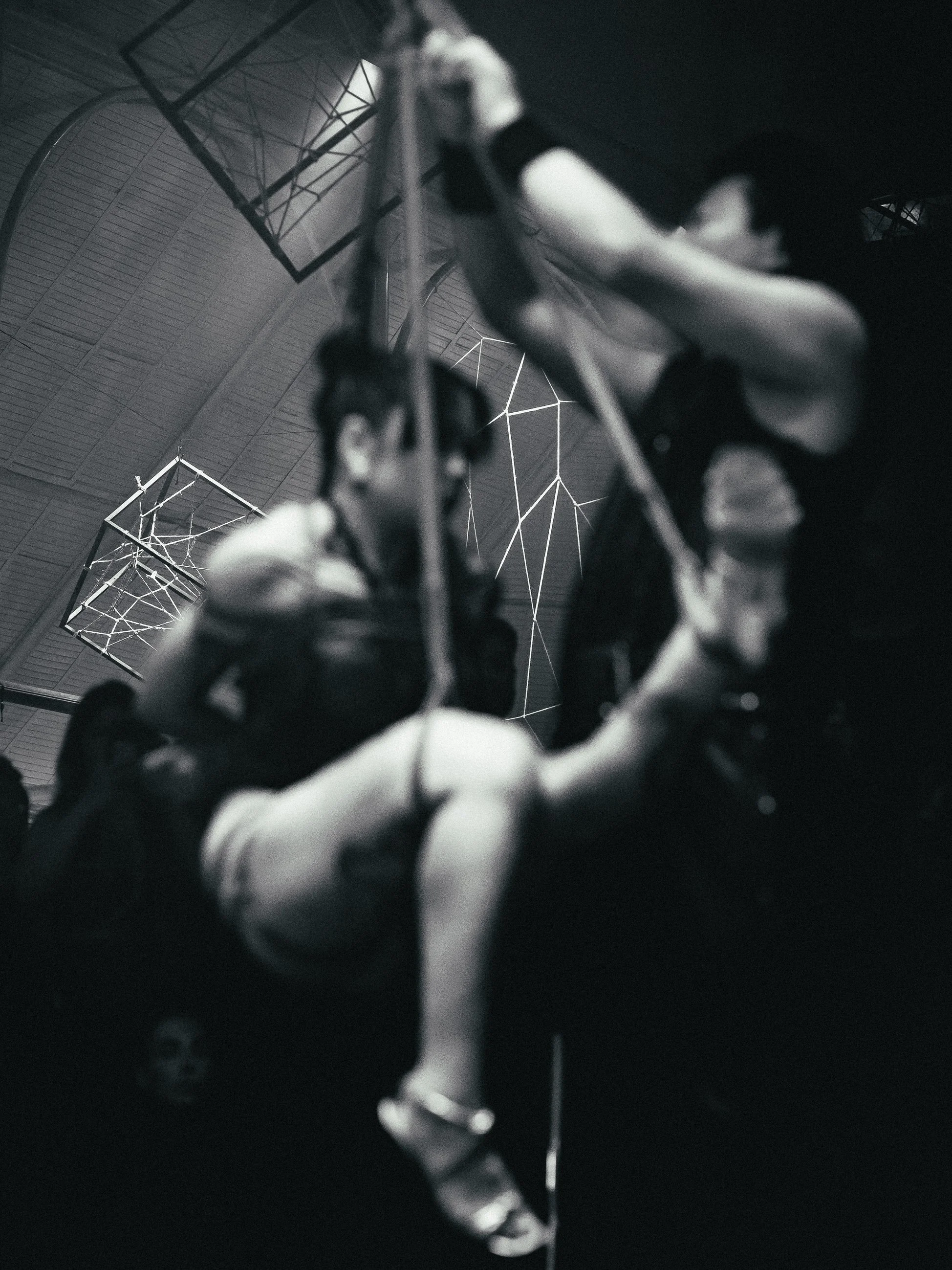 A person climbing or hanging on a rope or harness inside a dark, enclosed space with geometric light fixtures on the ceiling.