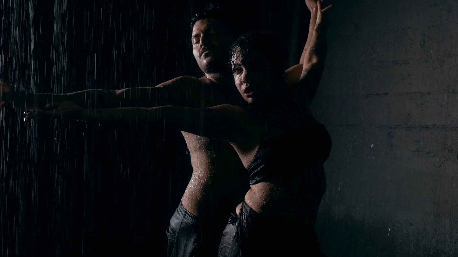 A man and woman in dark clothing are dancing closely together under rain against a dark background, with water droplets visible on their bodies.