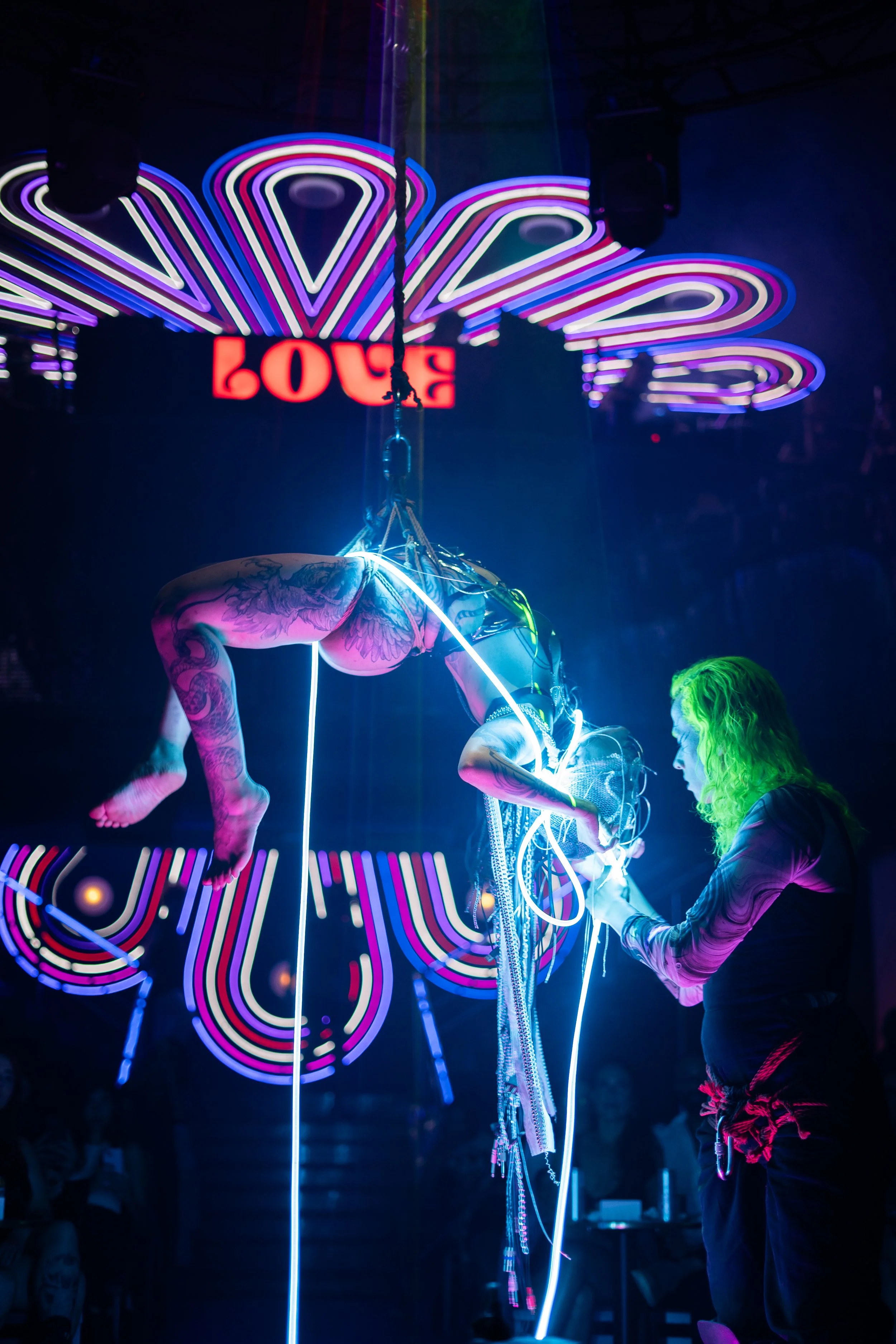 A performer with tattoos balancing on a vertical pole during a neon-lit circus act, with a performer supporting them and a bright, colorful sign reading 'LOVE' in the background.