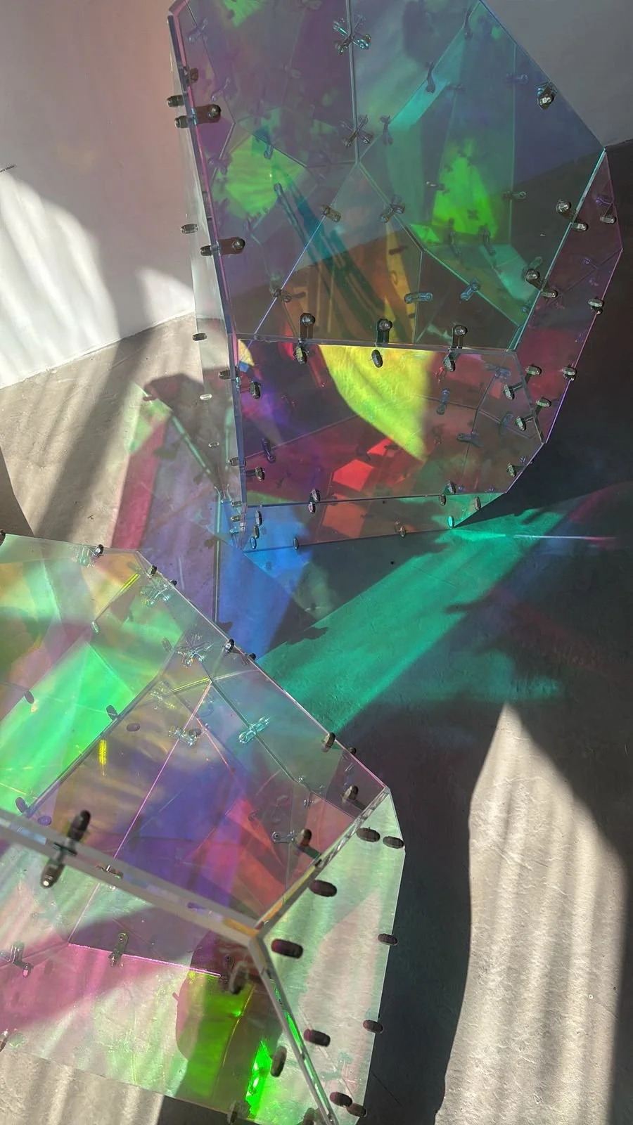 Two transparent, iridescent geometric sculptures reflecting colorful light shadows on the floor.
