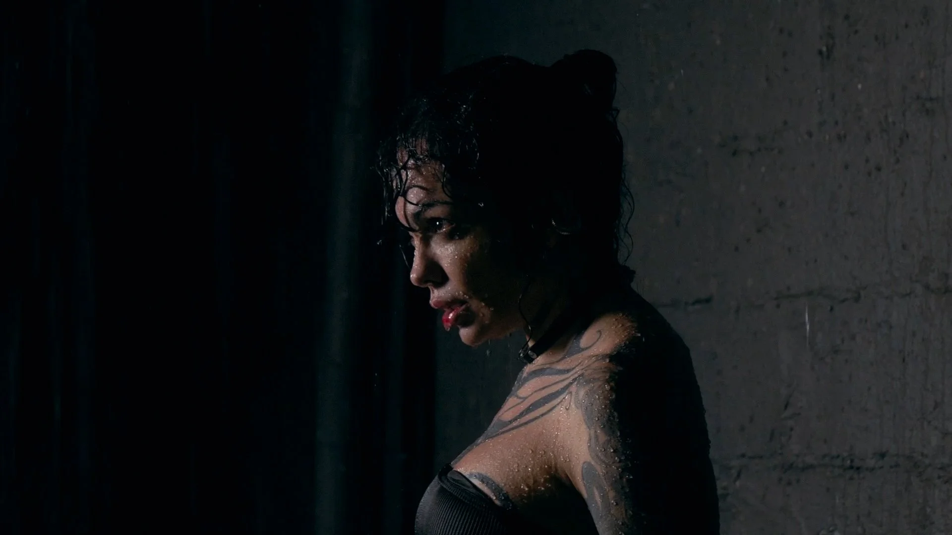 A woman with wet hair and tattoos on her shoulder and arm, looking down and to the side, in a dark setting with rain or water droplets on her skin.