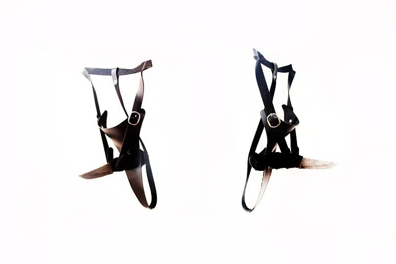 Two black leather harnesses with metal buckles, each with attached knives, arranged against a white background.