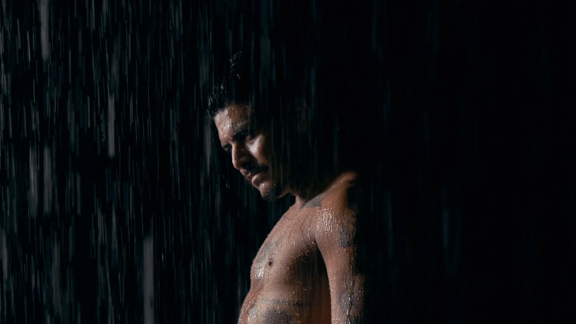 A shirtless man standing in the rain with his head bowed and eyes closed, illuminated against a dark background.